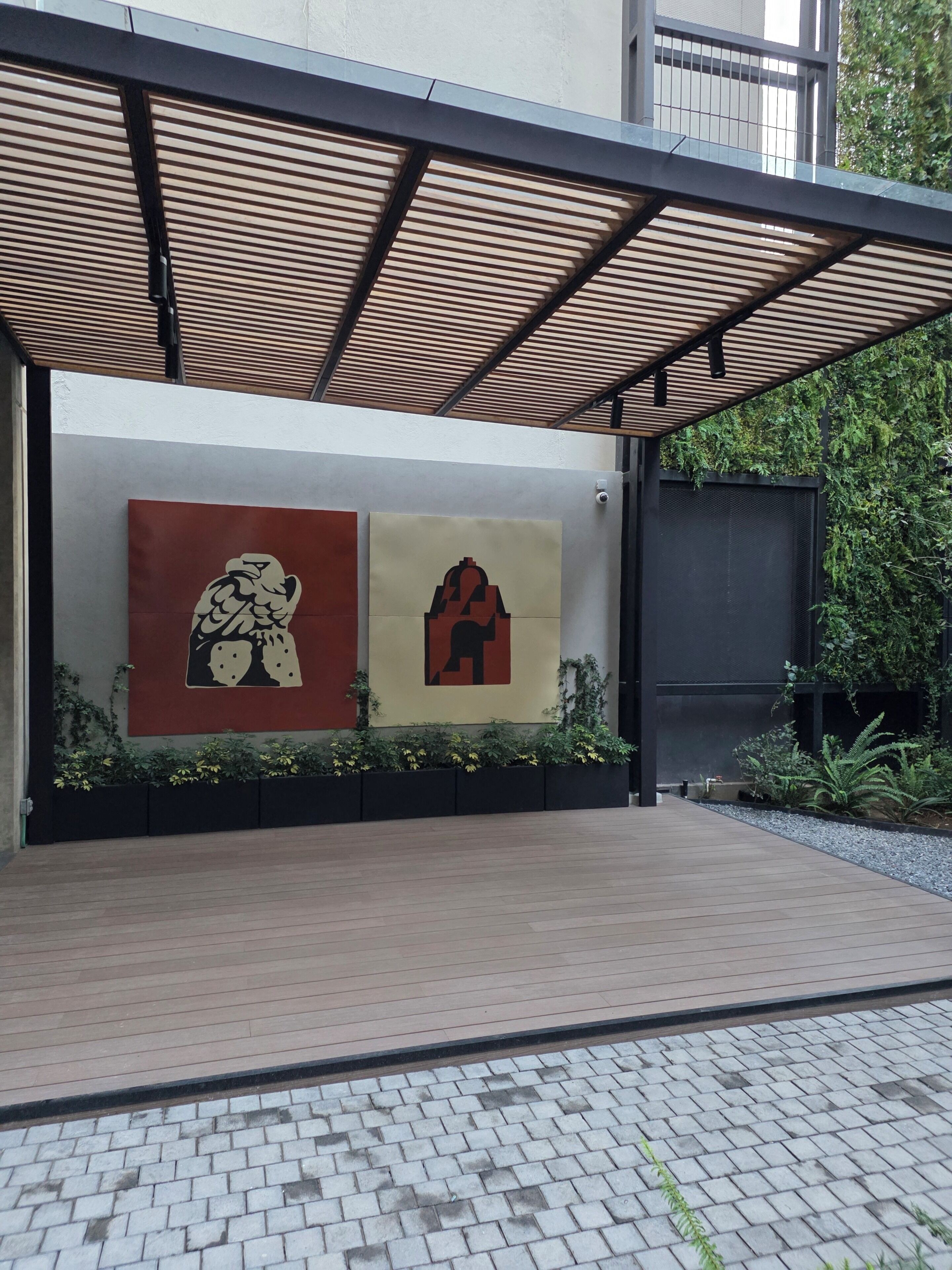 A covered outdoor area with two artworks on a wall, a paved walkway, and a garden with plants.