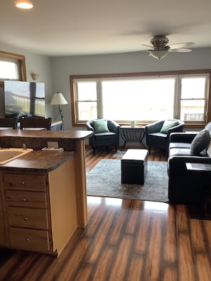 A spacious living area with a kitchen, large windows, and a ceiling fan.