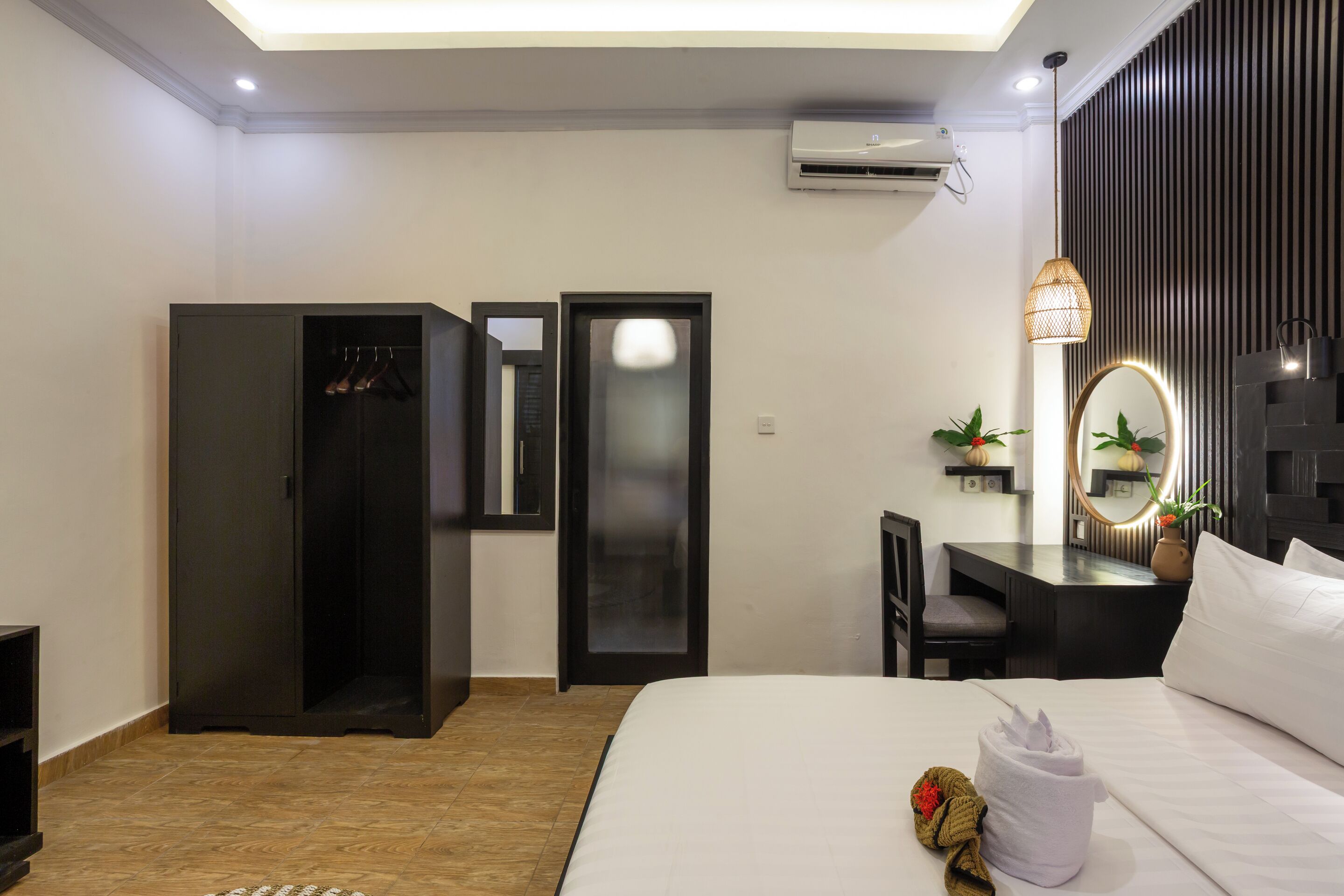 A hotel room with a bed, a wardrobe, a desk with a chair, a mirror, and an air conditioning unit.