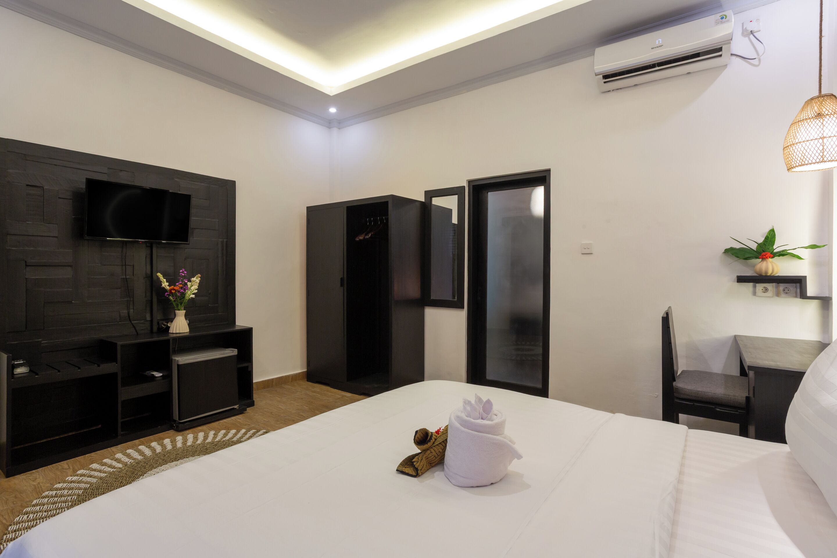 A modern hotel room with a flat-screen TV, a wardrobe, a desk with a chair, and a bed with a towel and slippers.