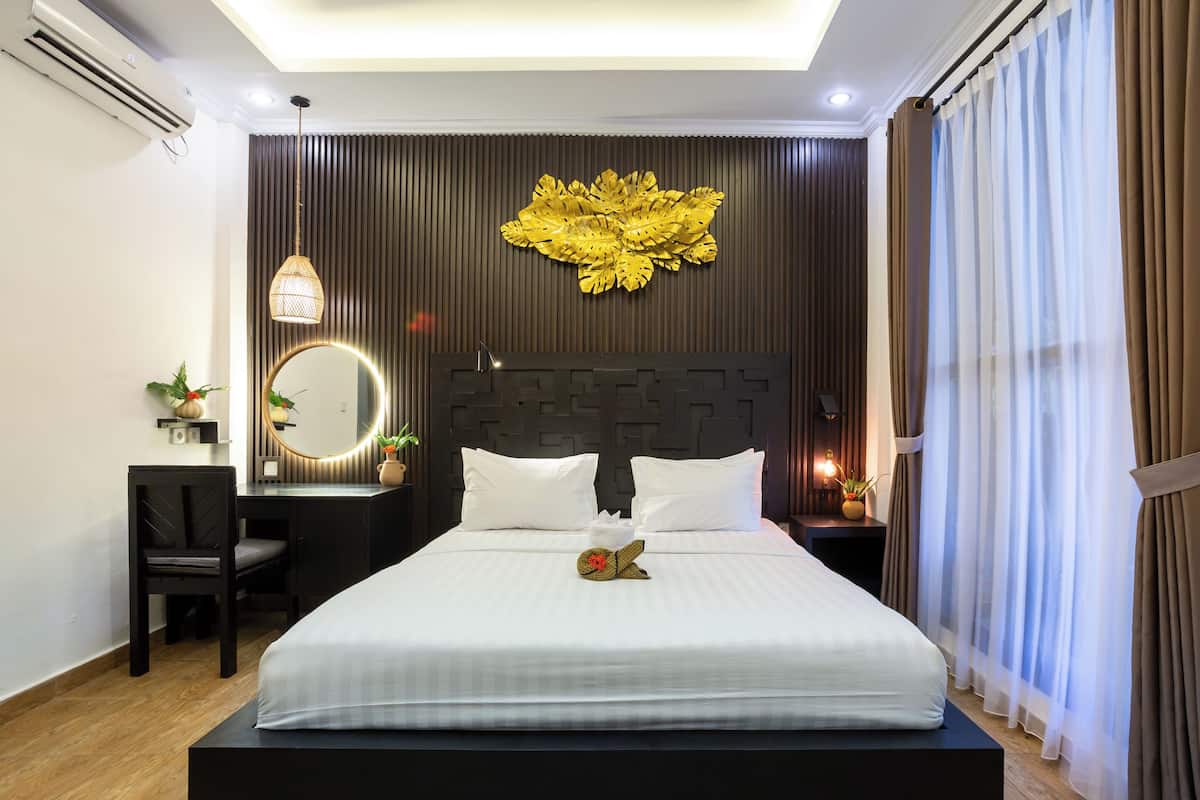 Habitación de hotel con cama, escritorio, silla, espejo, un adorno en la pared y mesita de noche con lámpara.