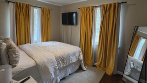 Un dormitorio con cama, televisor de pantalla plana, mesita de noche con lámpara y pañuelos, y cortinas amarillas.