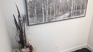 A framed black and white forest photograph on a wall, a vase with dried plants, and a black rug on the floor.