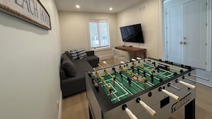 A room with a foosball table, a sofa, a TV, and a door. - Beachfront Luxury Lake Erie *Sleeps 14* HOT TUB. Great for Hosting Groups (Dunnville)