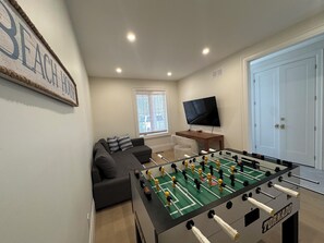 A room with a foosball table, a sofa, a TV, and a door. - Beachfront Luxury Lake Erie *Sleeps 14* HOT TUB. Great for Hosting Groups (Dunnville)