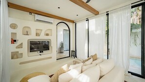 Een moderne woonkamer met een grote flatscreen-tv, een beige bank en een zwembad dat zichtbaar is door de glazen deuren.