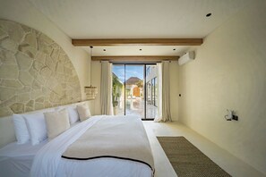 A bedroom with a stone wall, a large bed, a sliding glass door leading to a balcony, and a ceiling-mounted air conditioning unit. - Casa Degaz Canggu (Canggu)
