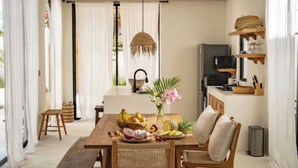 Een eetgedeelte met een houten tafel, rieten stoelen, een schaal met fruit en een hanglamp.