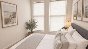 A bedroom with a large bed, two windows with blinds, a bedside table with a lamp, and framed pictures on the wall. - Luxury Country Feel Villa Retreat | Designer Home with elegant features (Wilton)