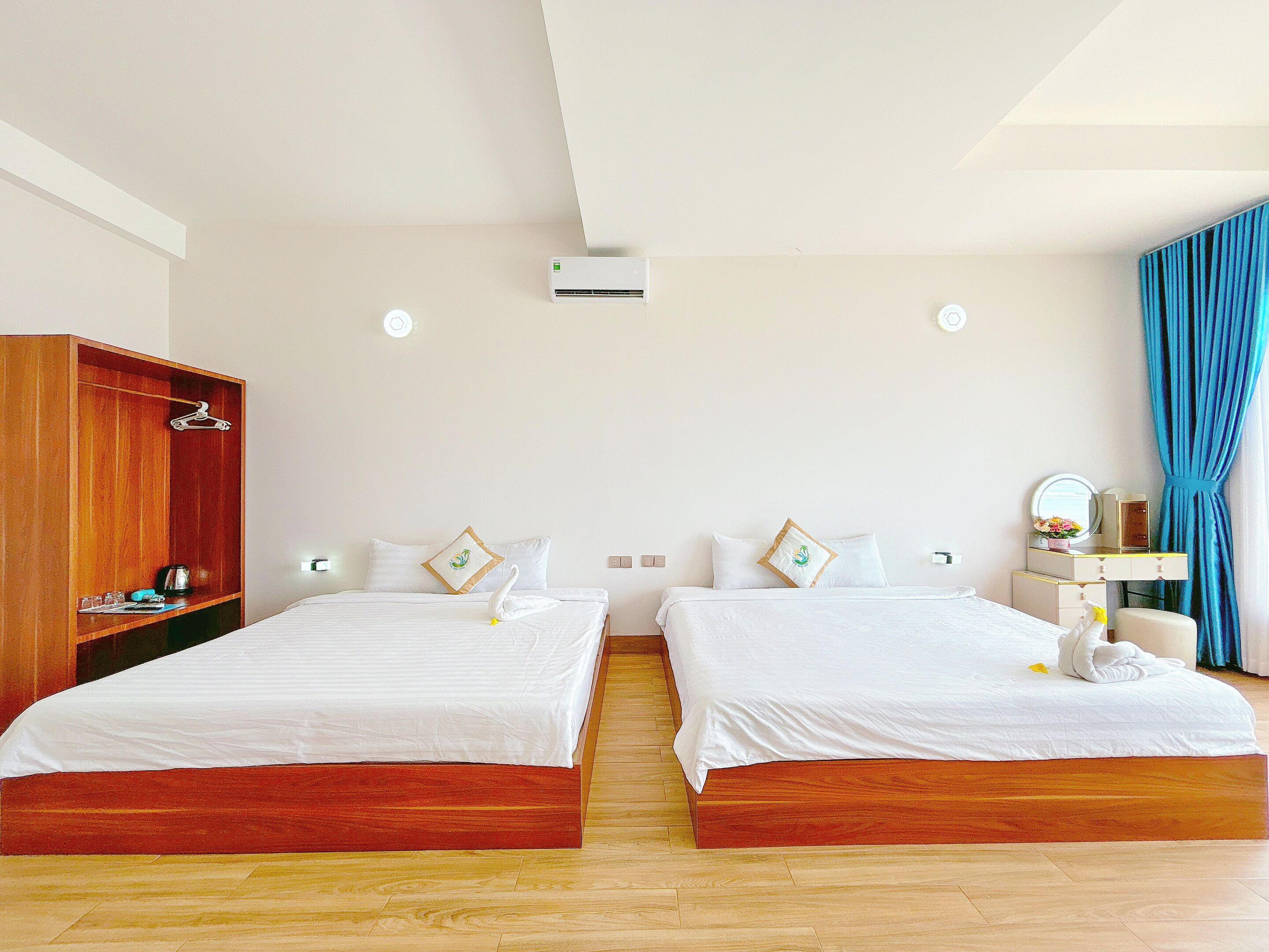 Een hotelkamer met twee bedden, een houten garderobe, een kaptafel met spiegel en een blauw gordijn.