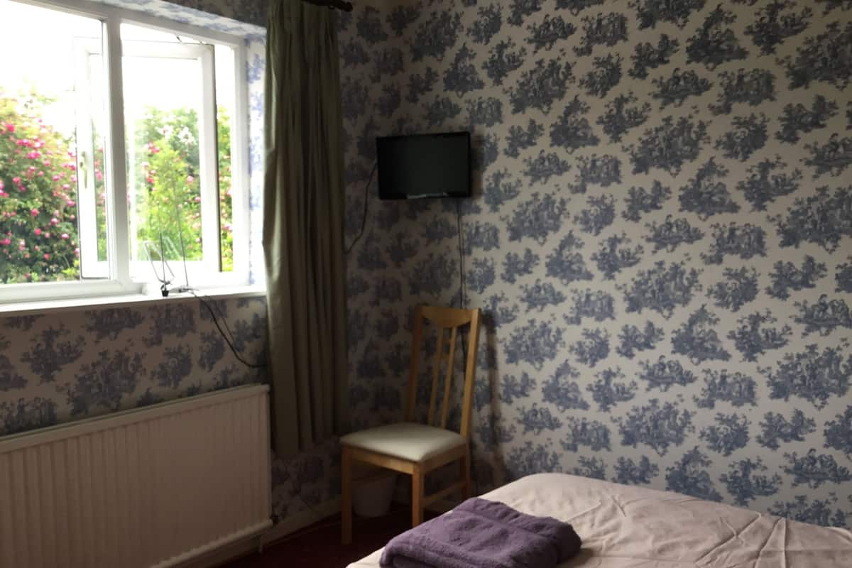 A room with a bed, a chair, a window with curtains, a radiator, and a television on the wall.