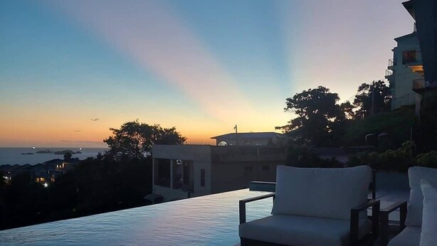An outdoor pool area with a view of the sunset, a lounge chair, and a table with a bottle and glasses. - Luxury 3 BR, Ocean view, With Pool, 15 mins from POS (Carenage)