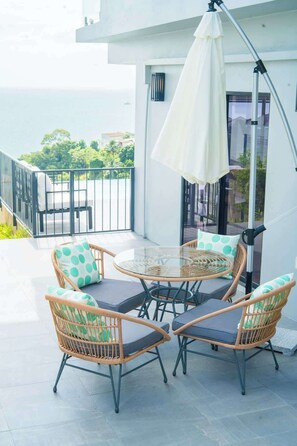 Ein Balkon mit Korbstühlen, einem Glastisch, einem weißen Sonnenschirm und Blick aufs Meer.