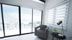 A modern room with large windows, a grey armchair, a small table with a lamp, and a plant. - Luxury 3 BR, Ocean view, With Pool, 15 mins from POS (Carenage)