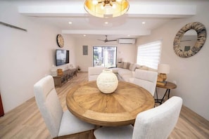 Ein Essbereich mit einem runden Holztisch, weißen Polsterstühlen, einer Blumenvase als Mittelstück, einem Deckenventilator und einem Fernseher an der Wand.