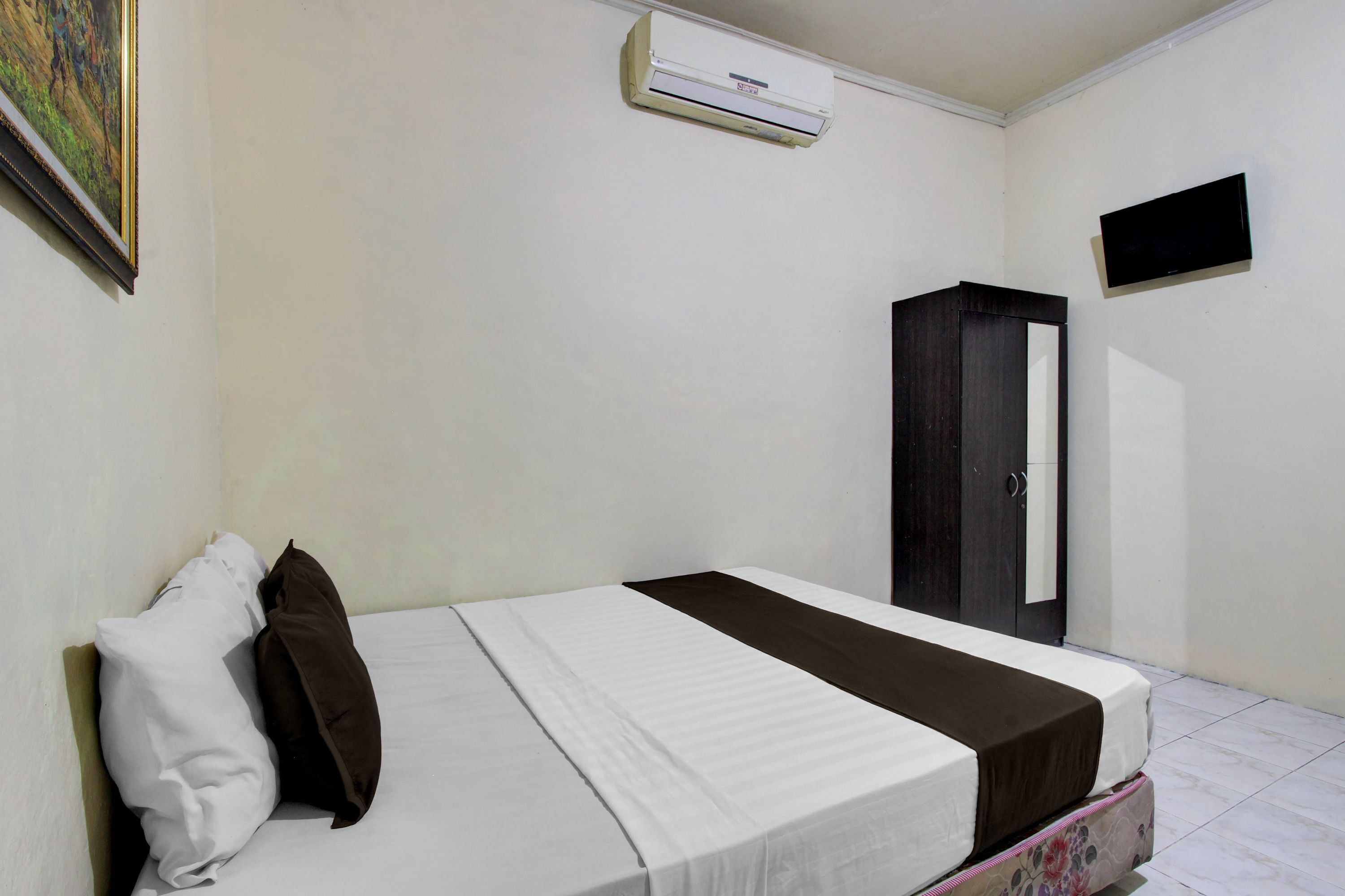 A single bed with a white and brown bedspread, a white pillow, a dark-colored wardrobe, a flat-screen TV mounted on the wall, and an air conditioning unit above the bed.