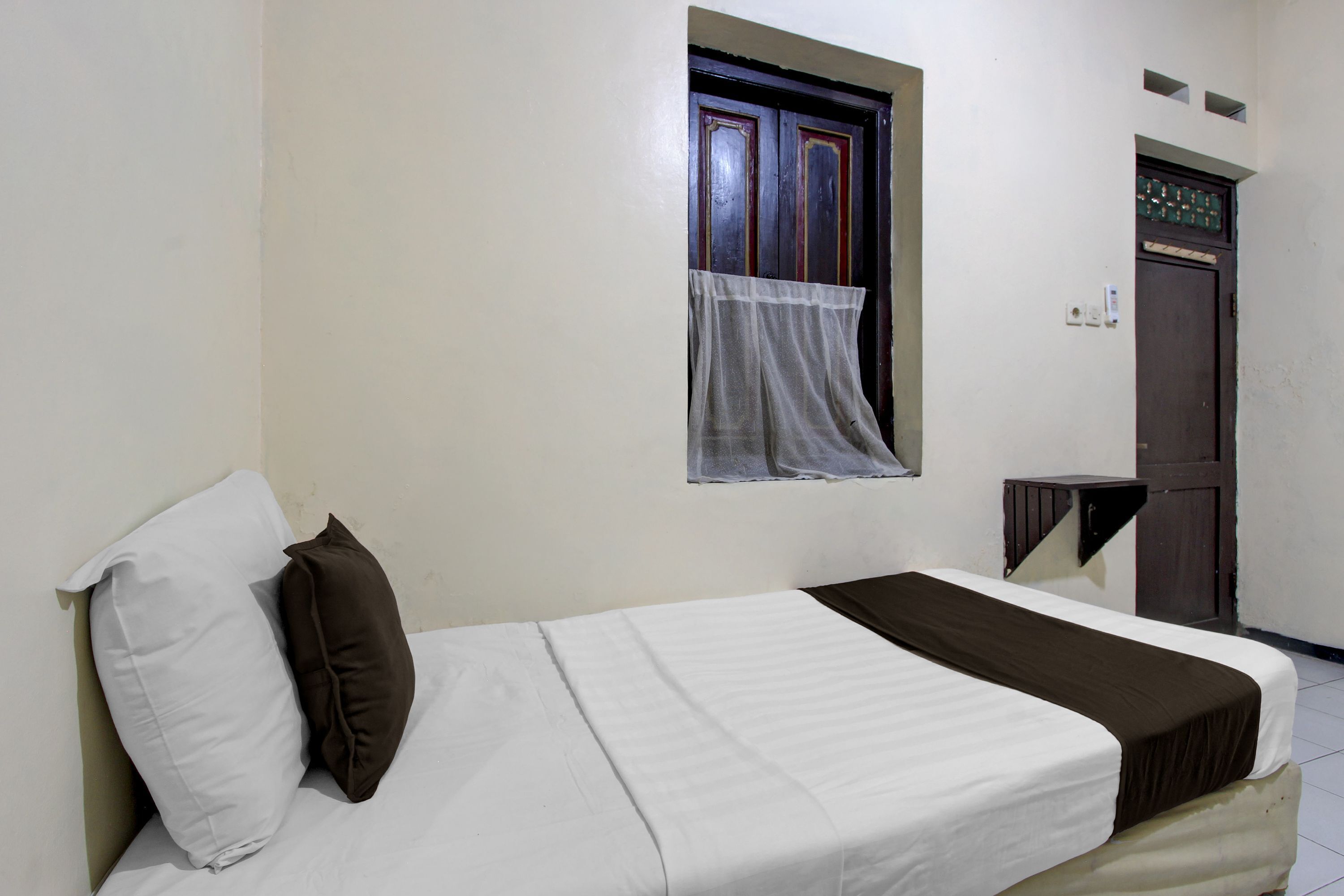 A single bed with a white and brown bedspread, a white pillow, and a brown pillow. There is a window with a sheer curtain and a wooden door with a decorative panel.