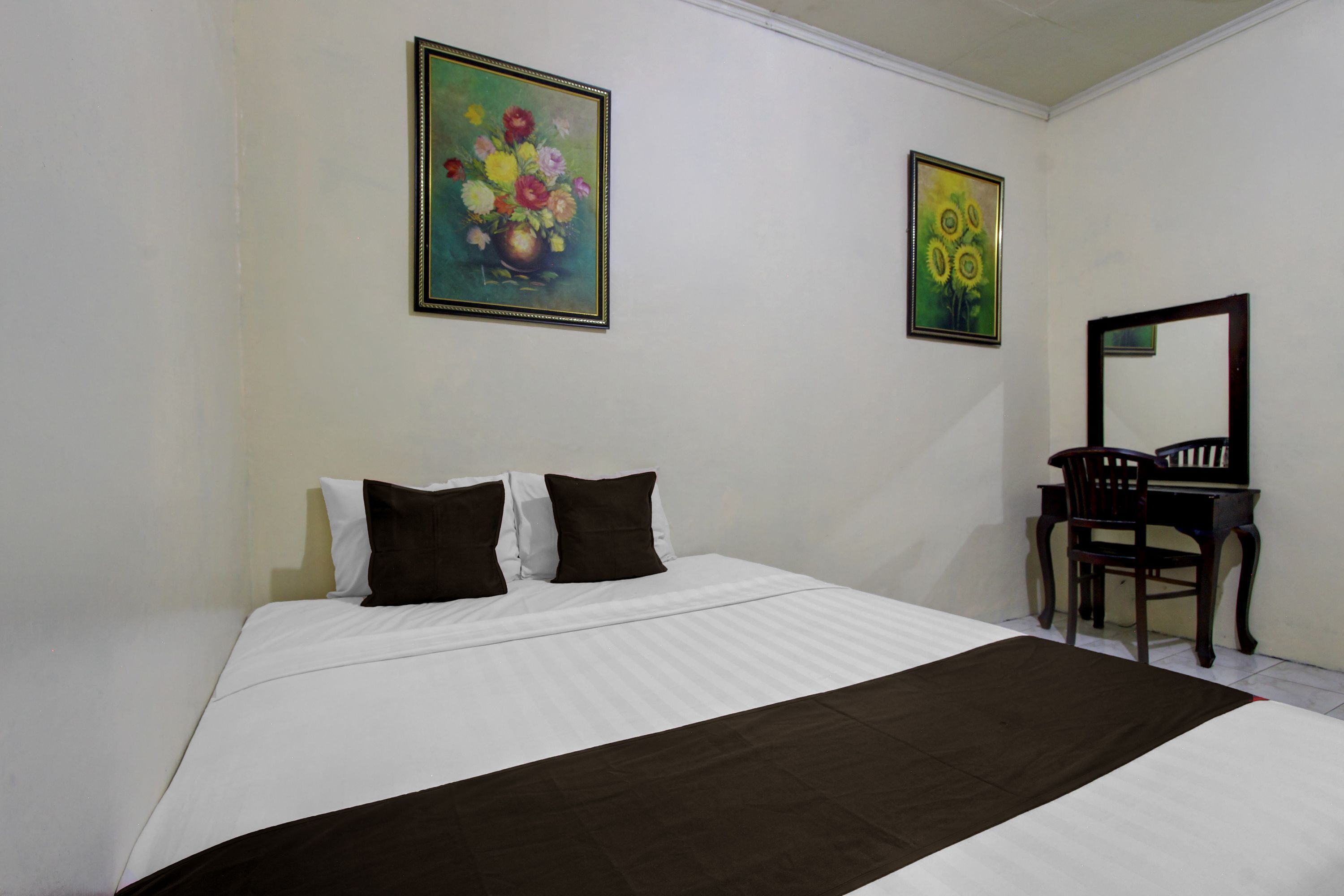 Uma cama bem arrumada com roupa de cama branca e castanha, uma cadeira de madeira e duas pinturas emolduradas na parede.