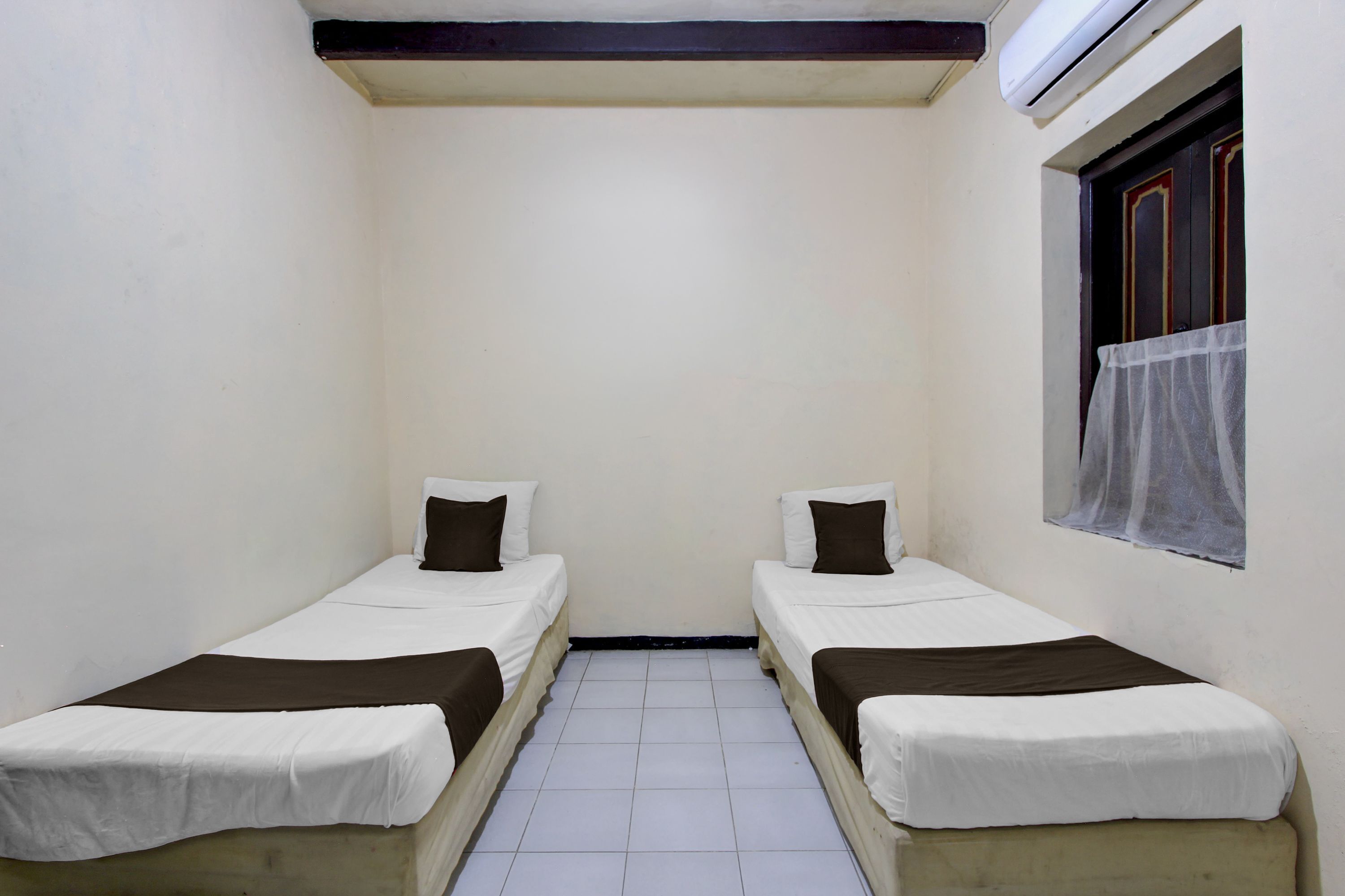 A small, clean room with two single beds, a window with a curtain, and a ceiling-mounted air conditioner.