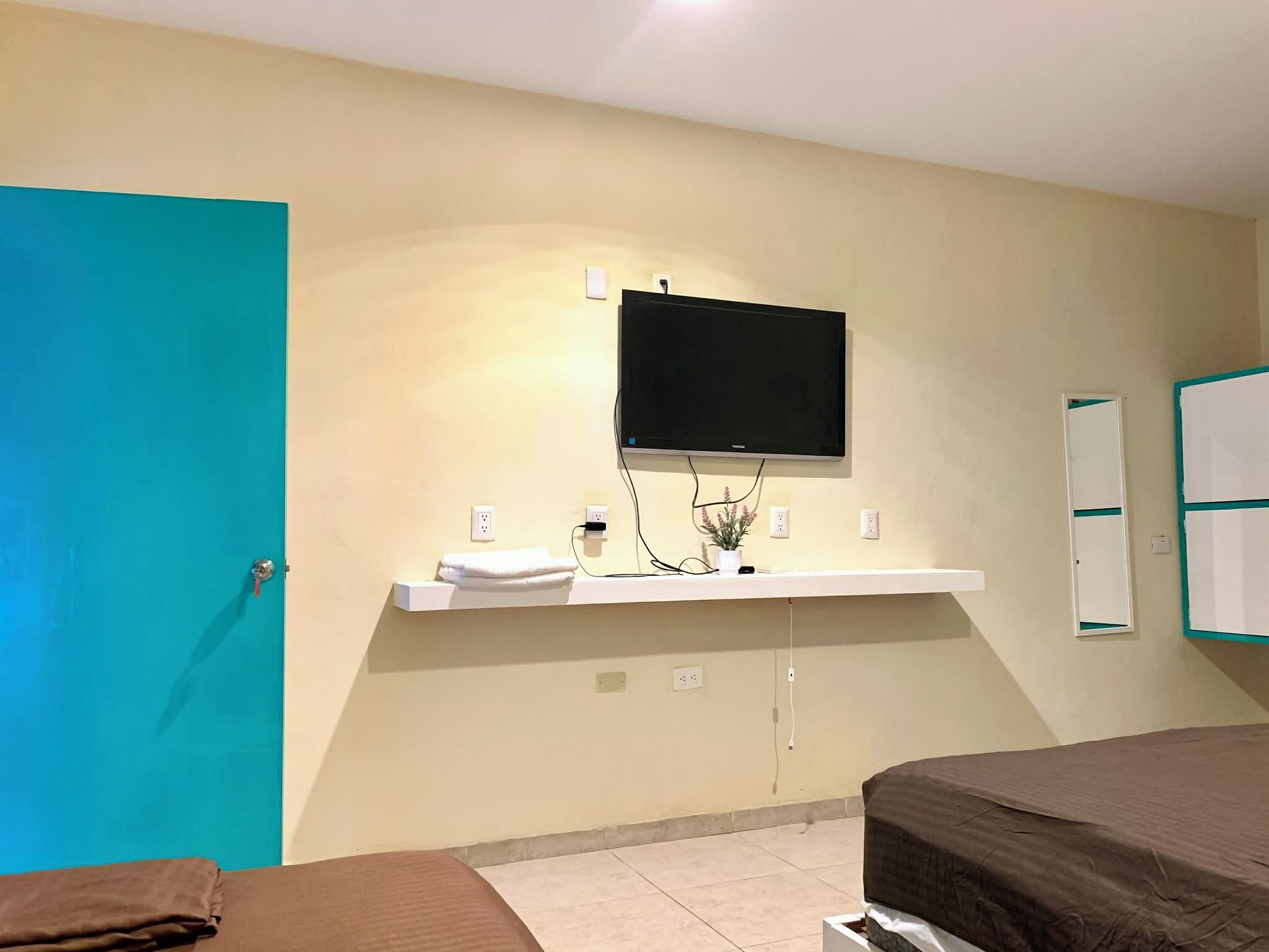 A hotel room with a teal door, a mounted TV, a white shelf with a towel, a small potted plant, and a bed with a brown cover.
