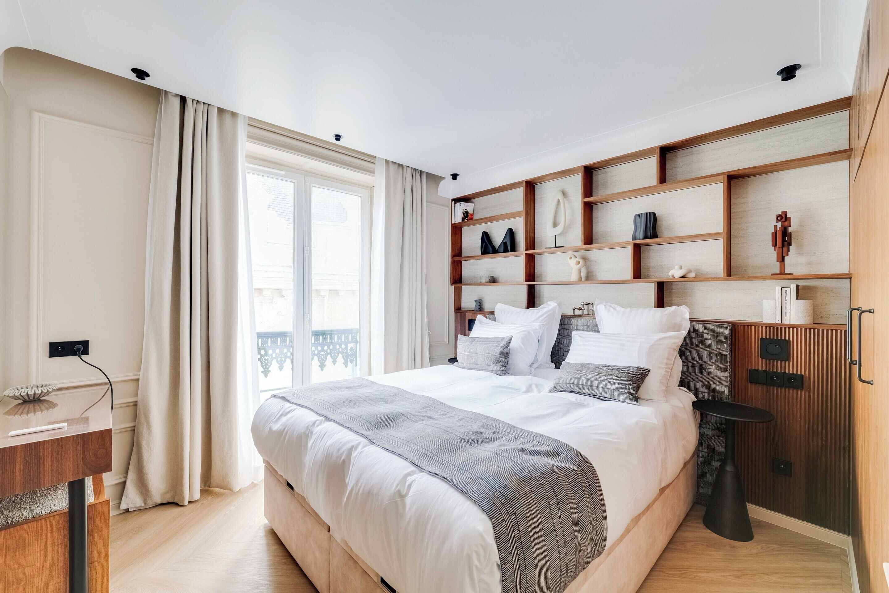 Quarto moderno com uma cama grande, uma mesinha de cabeceira, prateleiras embutidas e vista para o exterior.