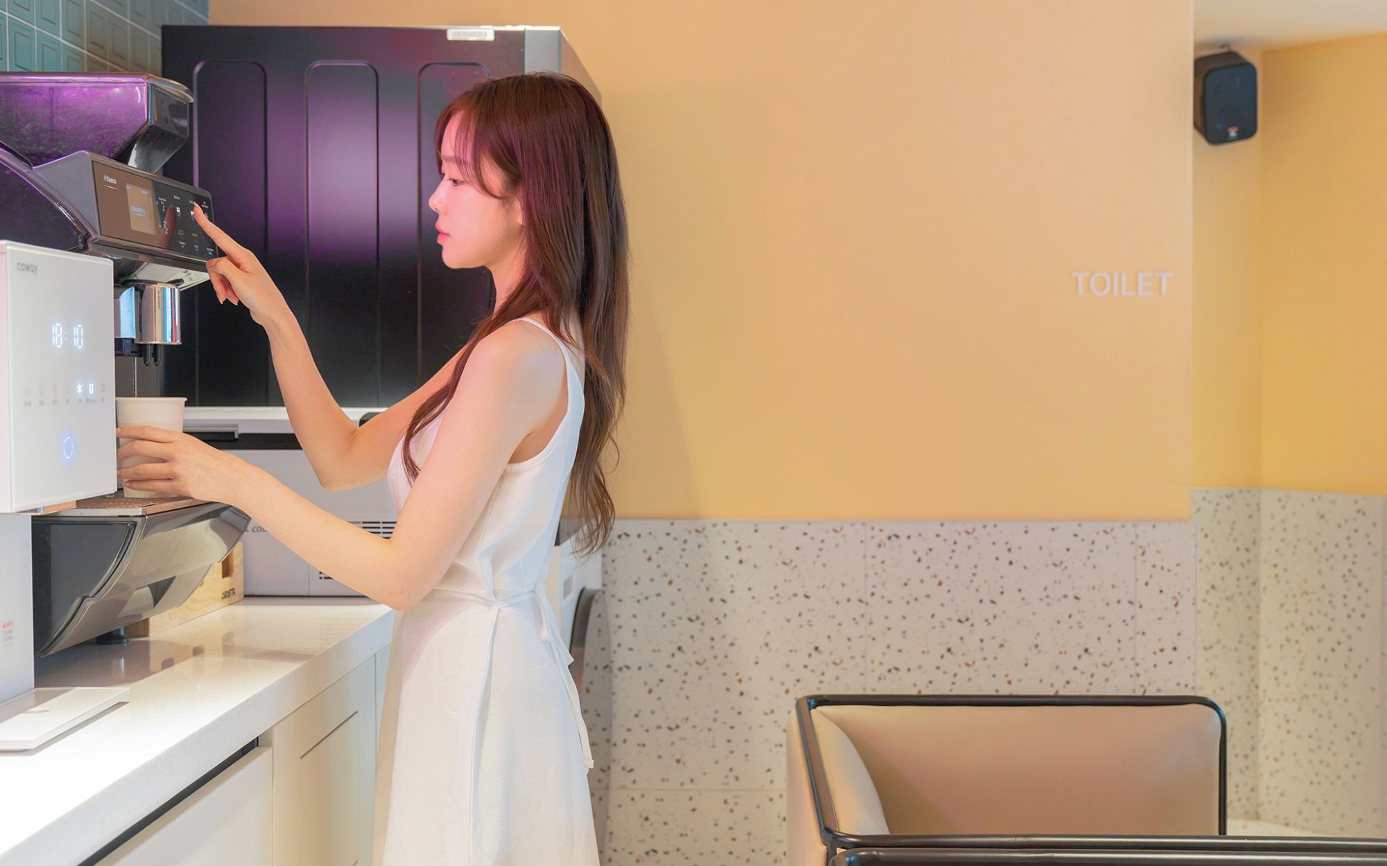 Een persoon in een witte jurk bedient een koffieapparaat in een moderne keuken met een zichtbaar toiletbord.