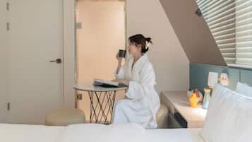 Une chambre d’hôtel avec un lit, une petite table ronde sur laquelle se trouvent un livre et une tasse, une table de chevet agrémentée d’objets décoratifs, et une porte donnant sur une autre pièce.