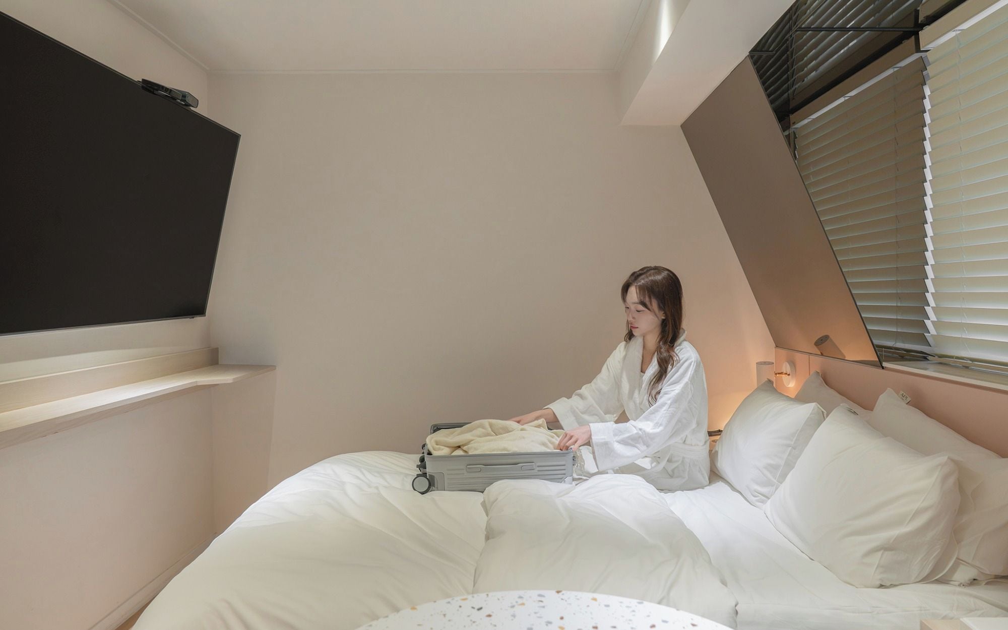 Een kamer met een bed, een raam met jaloezieën, een wandlamp en een verlaagd plafond.