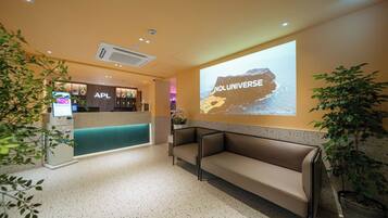 Un espace d’accueil d’hôtel comprenant un kiosque numérique, un comptoir d’accueil, un coin salon et un écran de projection diffusant l’image de « NOL UNIVERSE ».