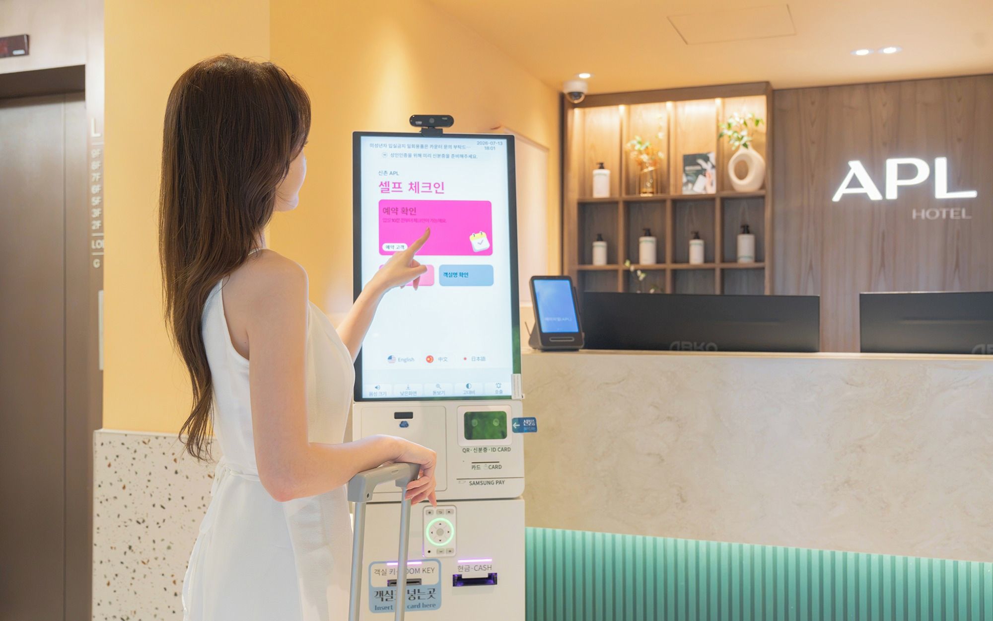 A person using a digital kiosk in a hotel lobby with a reception desk and a sign that reads 'APL HOTEL'.