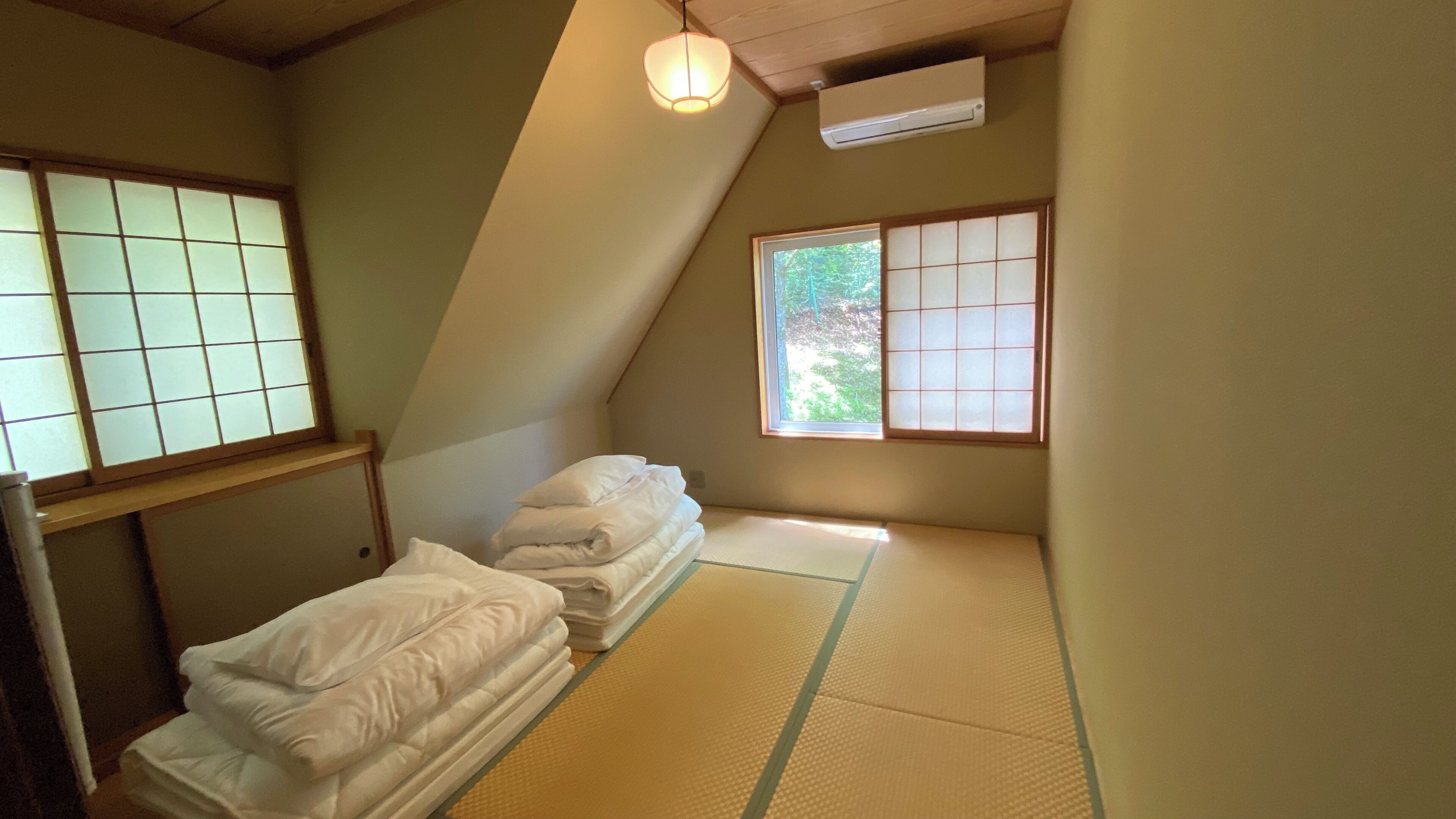 A room with tatami mats, a bed with white bedding, a window with a view of greenery, and a traditional Japanese-style air conditioning unit.