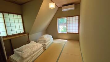 A room with tatami mats, a bed with white bedding, a window with a view of greenery, and a traditional Japanese-style air conditioning unit.