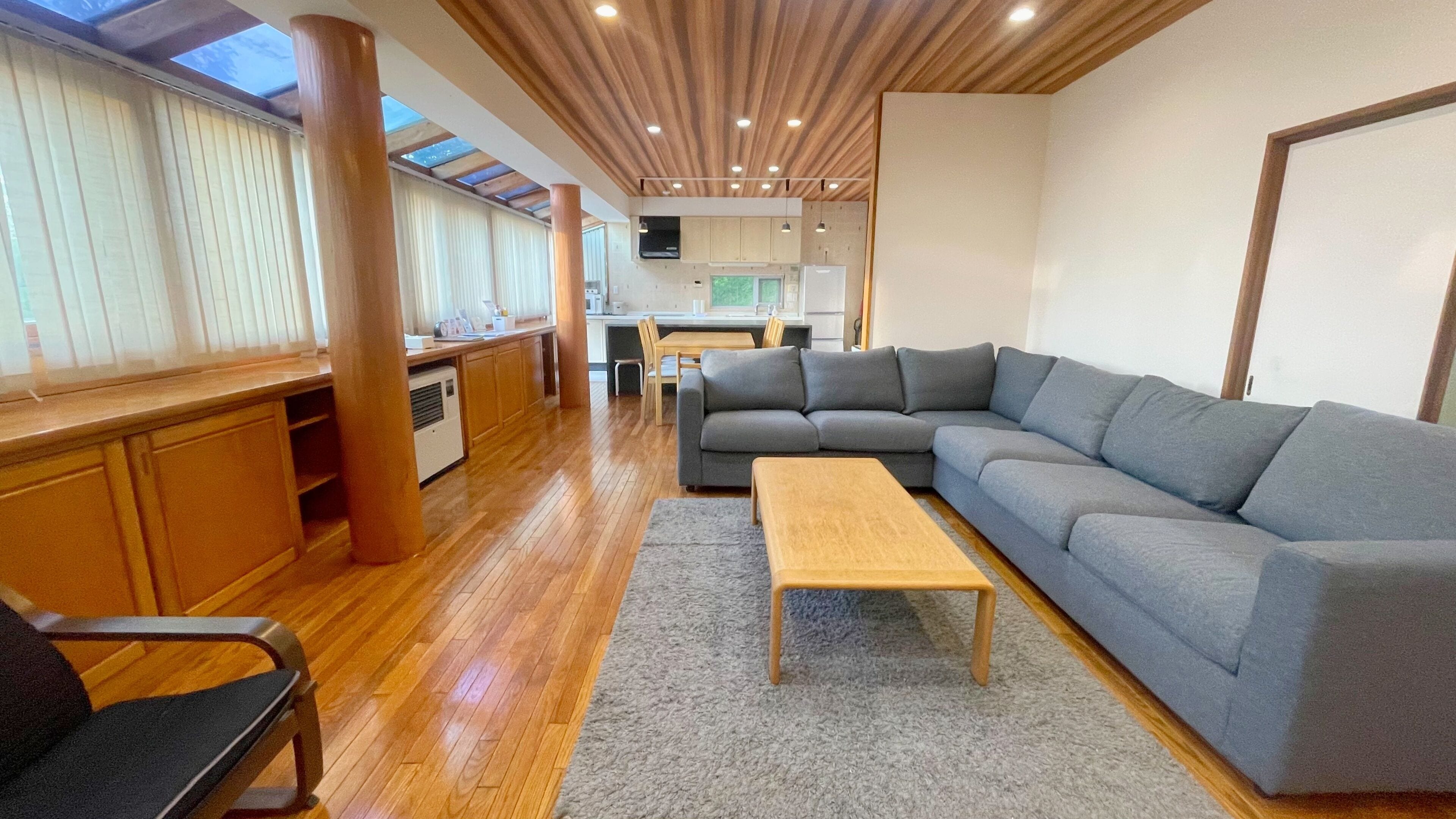 A modern interior with a wooden floor, a grey sofa, a wooden coffee table, and a kitchen area with a dining table and chairs.