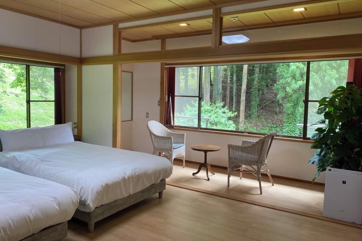 A bedroom with two beds, a small table, two chairs, and a large window with a forest view.