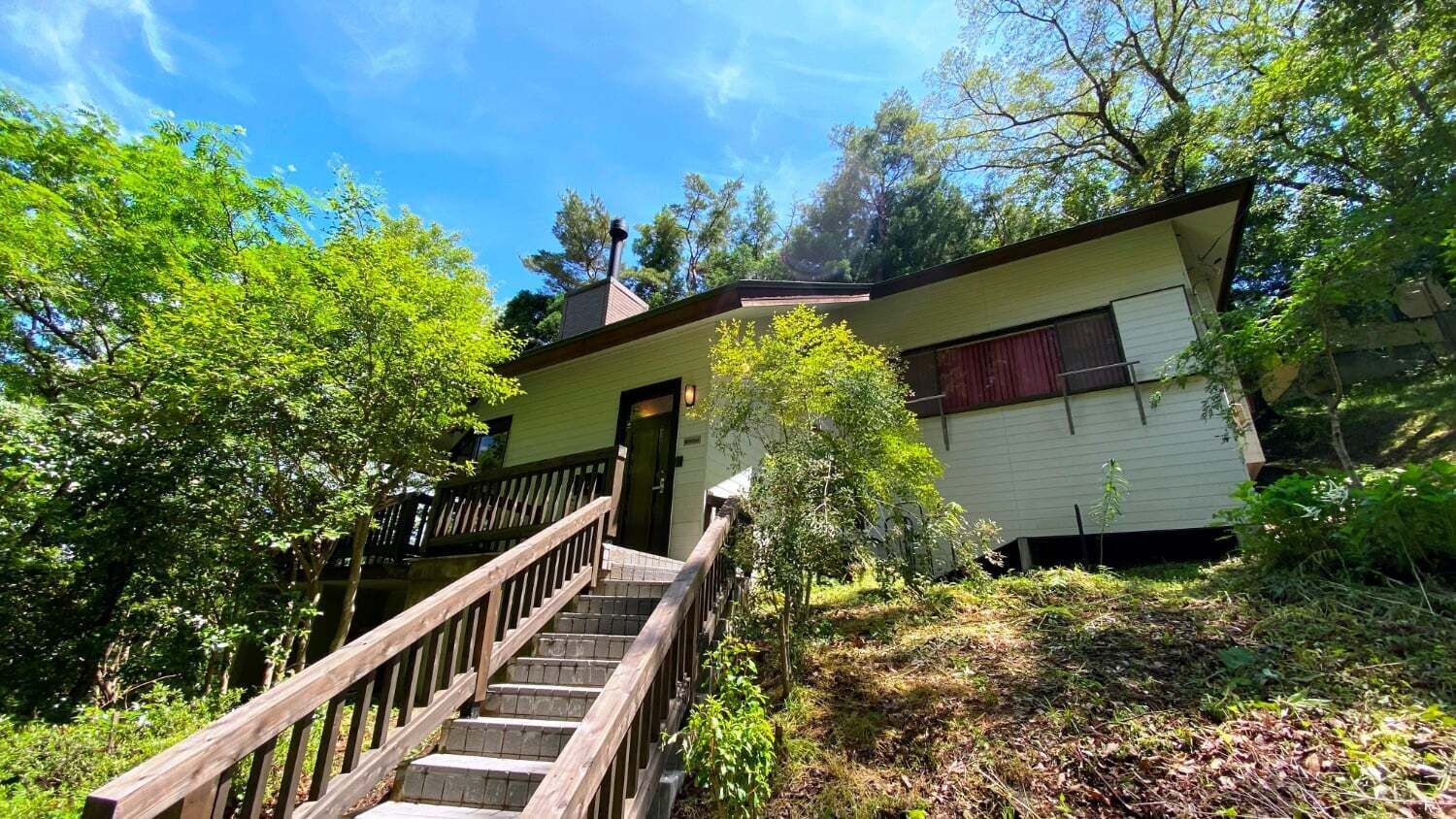 A two-story house with a wooden staircase leading to the front door, surrounded by dense greenery.
