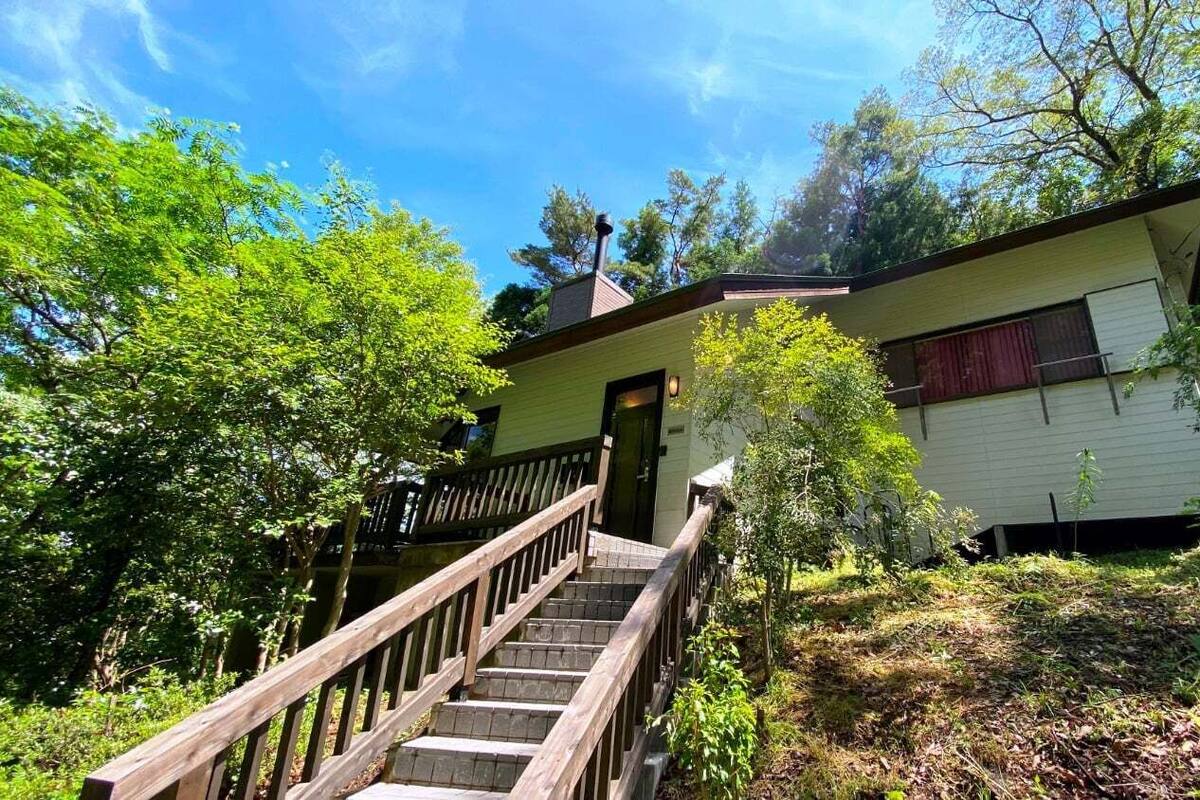 A two-story house with a wooden staircase leading to the front door, surrounded by dense greenery.