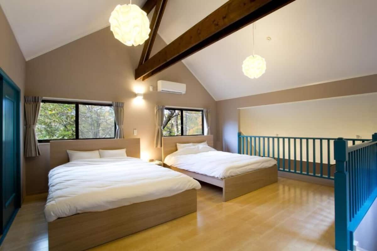 A bedroom with two beds, wooden flooring, a sloped ceiling with exposed beams, and large windows with curtains.