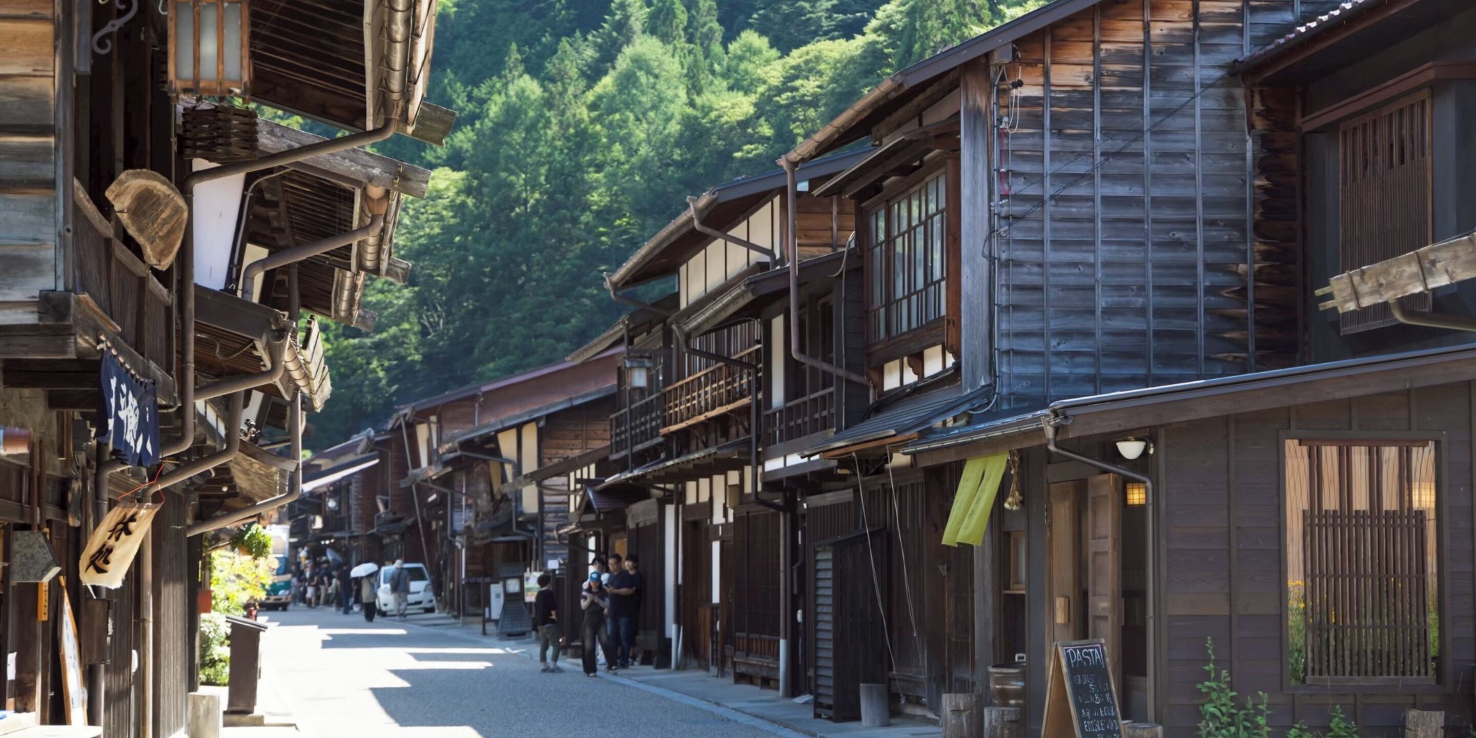 A traditional wooden building with a balcony, a street with pedestrians, and a forested hill in the background.