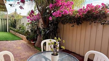 A round glass table with white plastic chairs, a potted plant with pink flowers, and a brick patio.