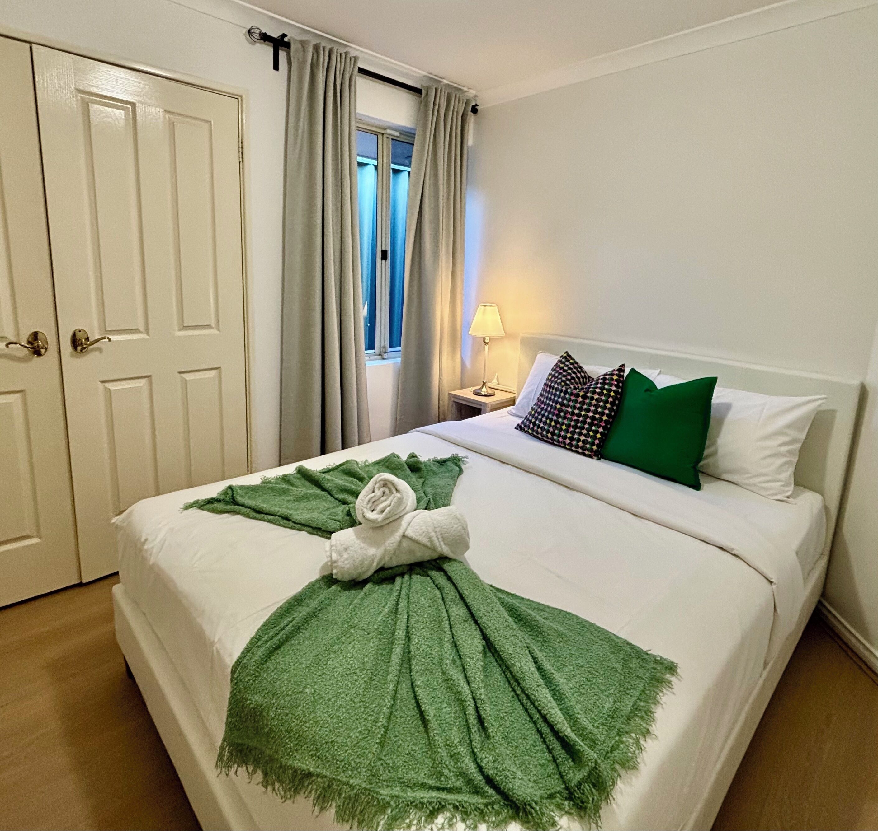 Een keurig opgemaakt bed met een groene handdoek en plaid, een lamp op het nachtkastje, en een raam met gordijnen.