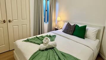 Een keurig opgemaakt bed met een groene handdoek en plaid, een lamp op het nachtkastje, en een raam met gordijnen.