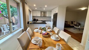 Un coin repas avec une table en bois dressée pour quatre personnes, des chaises blanches et une cuisine en arrière-plan.