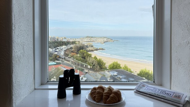 Ein Fenster mit Blick auf einen Strand, ein Fernglaspaar und eine Teller mit Croissants auf einem Fensterbrett.