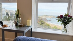 Ein Zimmer mit Bett, einem Tisch mit Kerze, Blumen in einer Vase und Blick auf Meer und Gebäude.