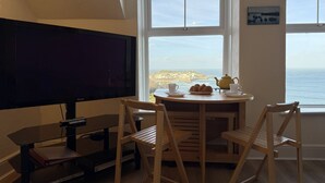 Ein Zimmer mit Fernseher, Tisch mit Stühlen, Fenster mit Meerblick und einer Uhr an der Wand.