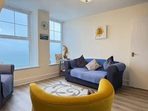 A living room with a blue sofa, a yellow armchair, a window with frosted glass, a wall clock, and a framed picture. - Draycott Dreams has stunning sea views across St Ives Bay & Harbour (St Ives)