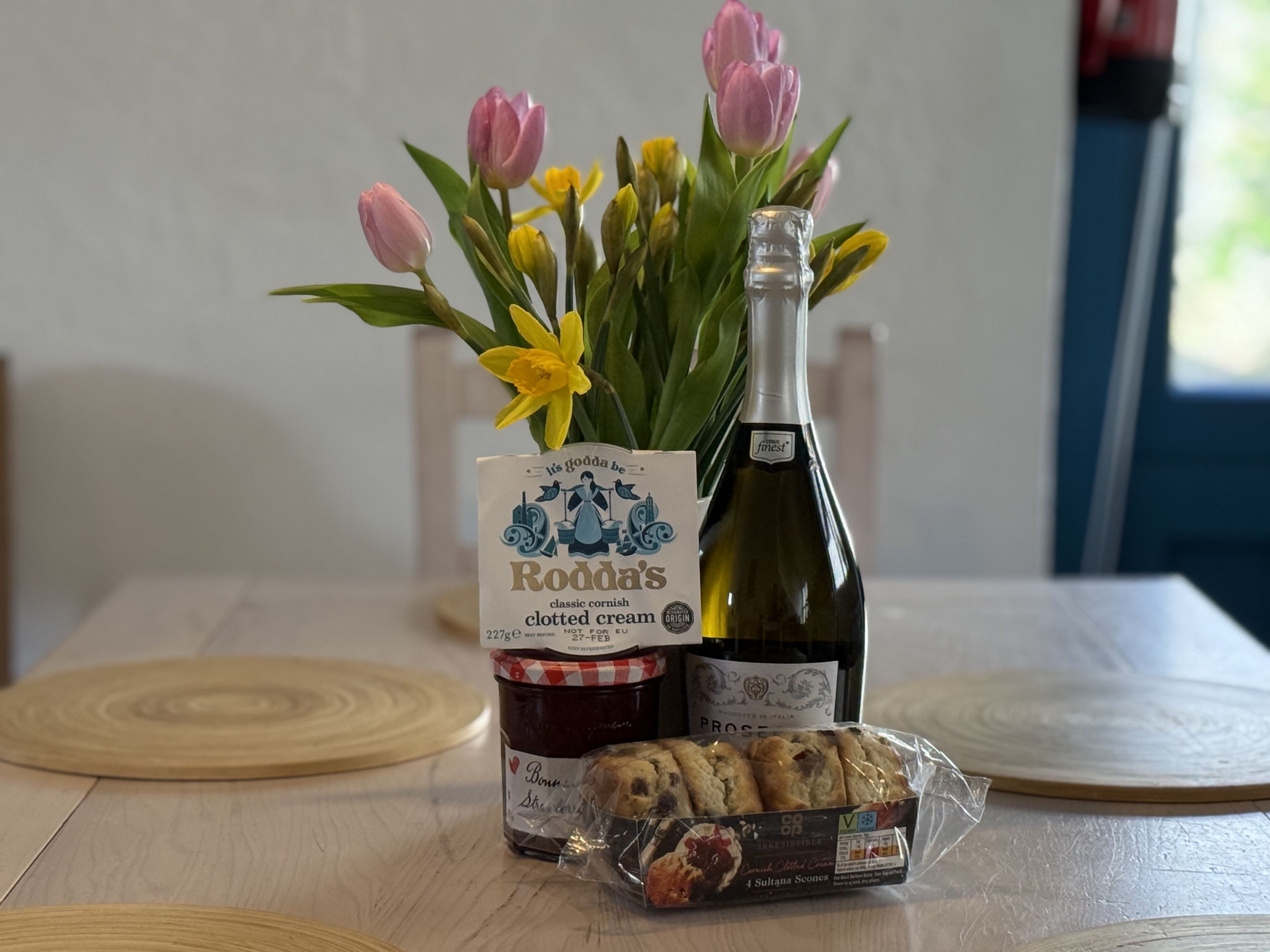 A table with a bottle of wine, a jar of clotted cream, a box of biscuits, and a bouquet of flowers.