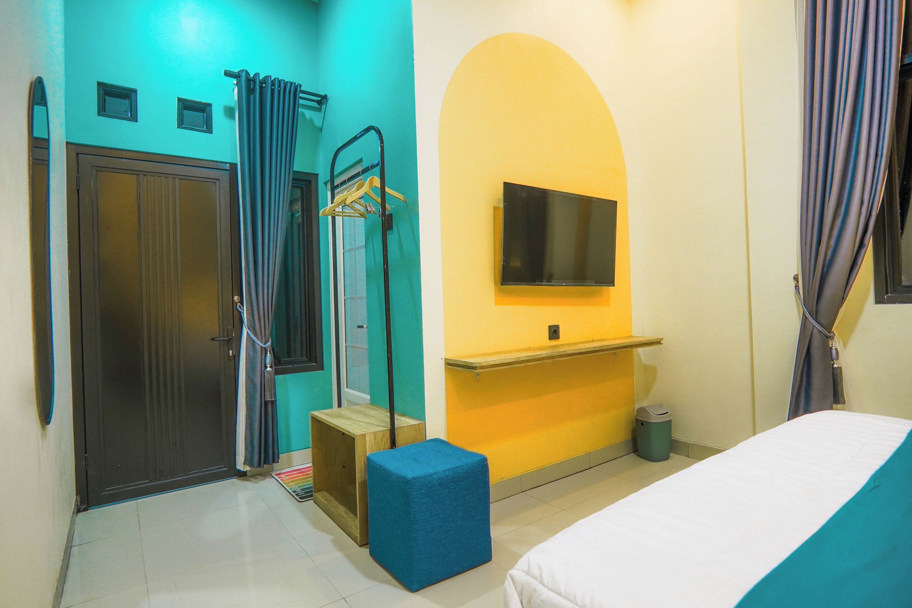 Una habitación con cama, televisor montado en la pared, puerta, zona de ducha y un taburete pequeño.