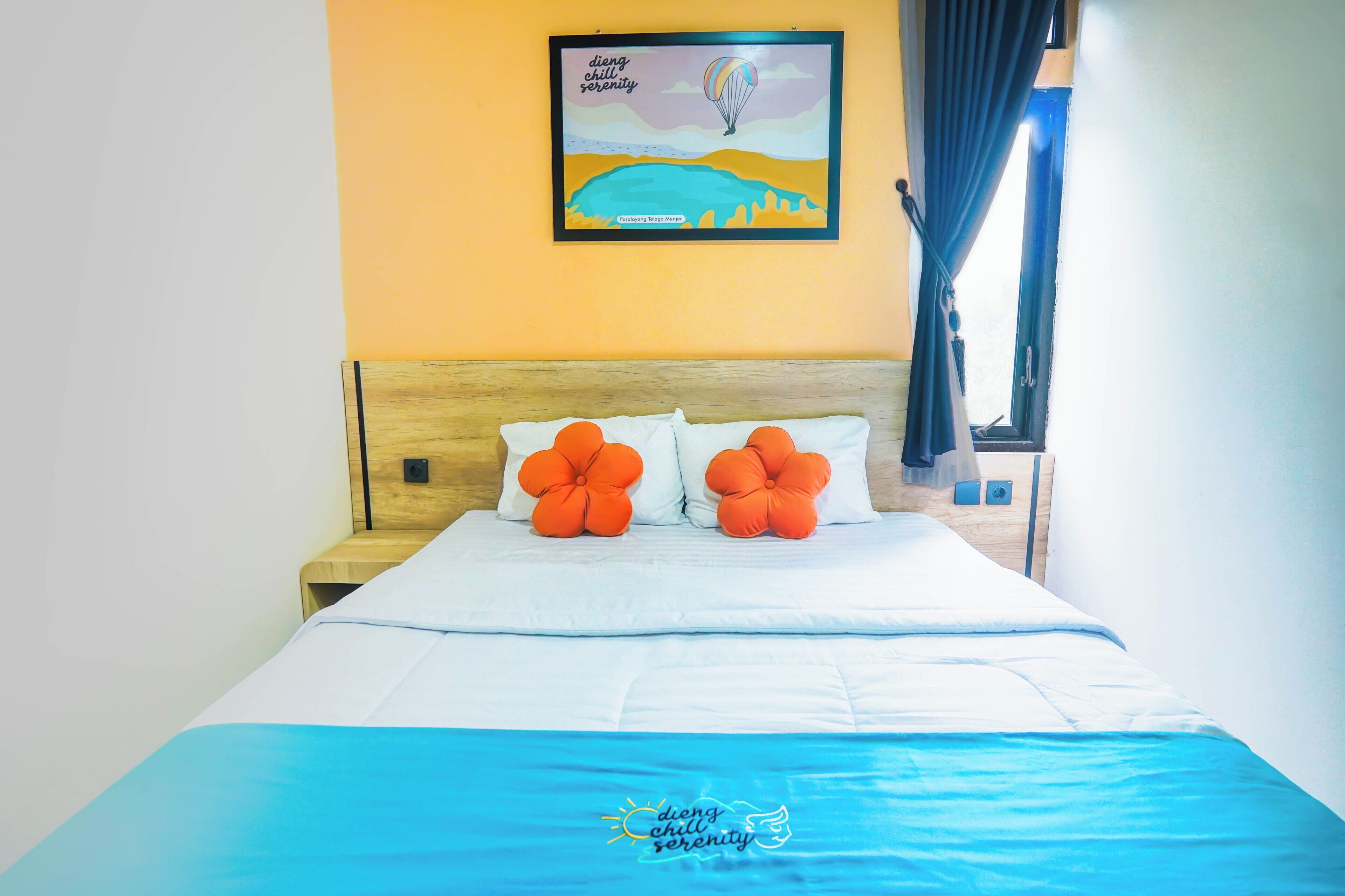 A bed with white bedding, two orange flower-shaped pillows, a framed picture on the wall, and a window with curtains.