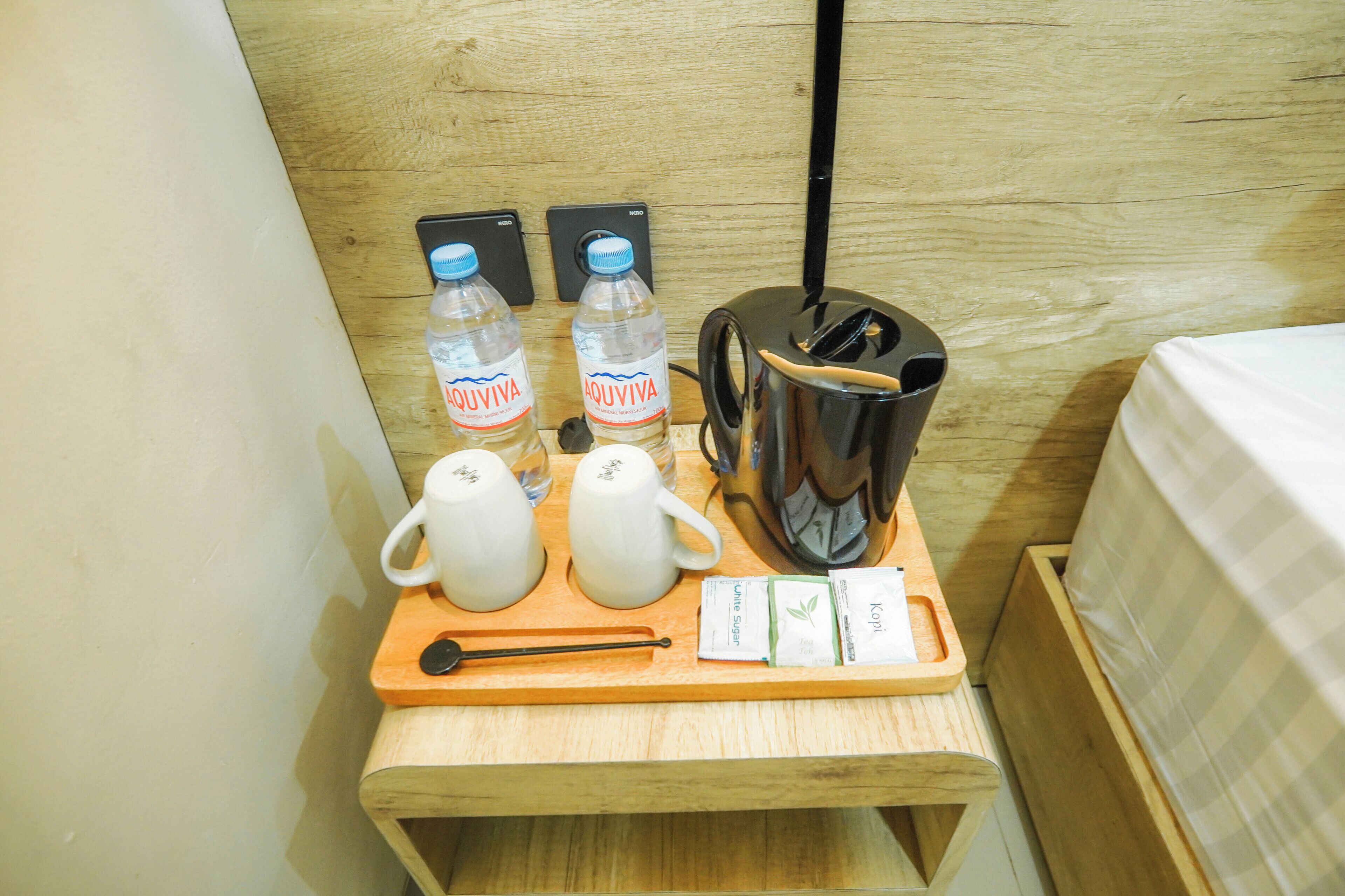 Una mesita de noche de madera con una tetera, dos botellas de agua, una taza, una cuchara y una bandeja con dos sobres de té.